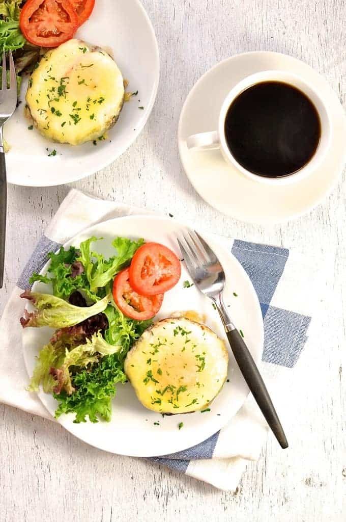 Overhead photo of Baked Eggs in Mushrooms covered in melted cheese on a white plate with coffee on the side, ready to be eaten.