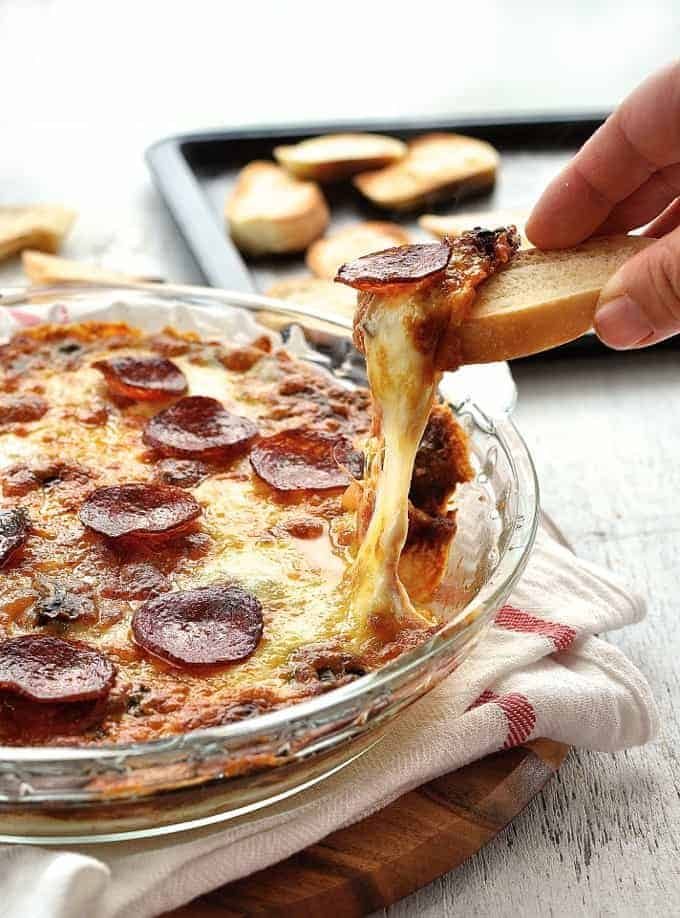 Toasted bread scooping up ultra cheesy, gooey Pizza Dip in a glass dish with a great cheese pull shot.