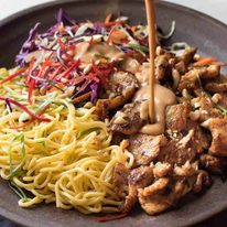 Peanut dressing being poured over Satay Chicken Noodle Salad in a brown bowl.