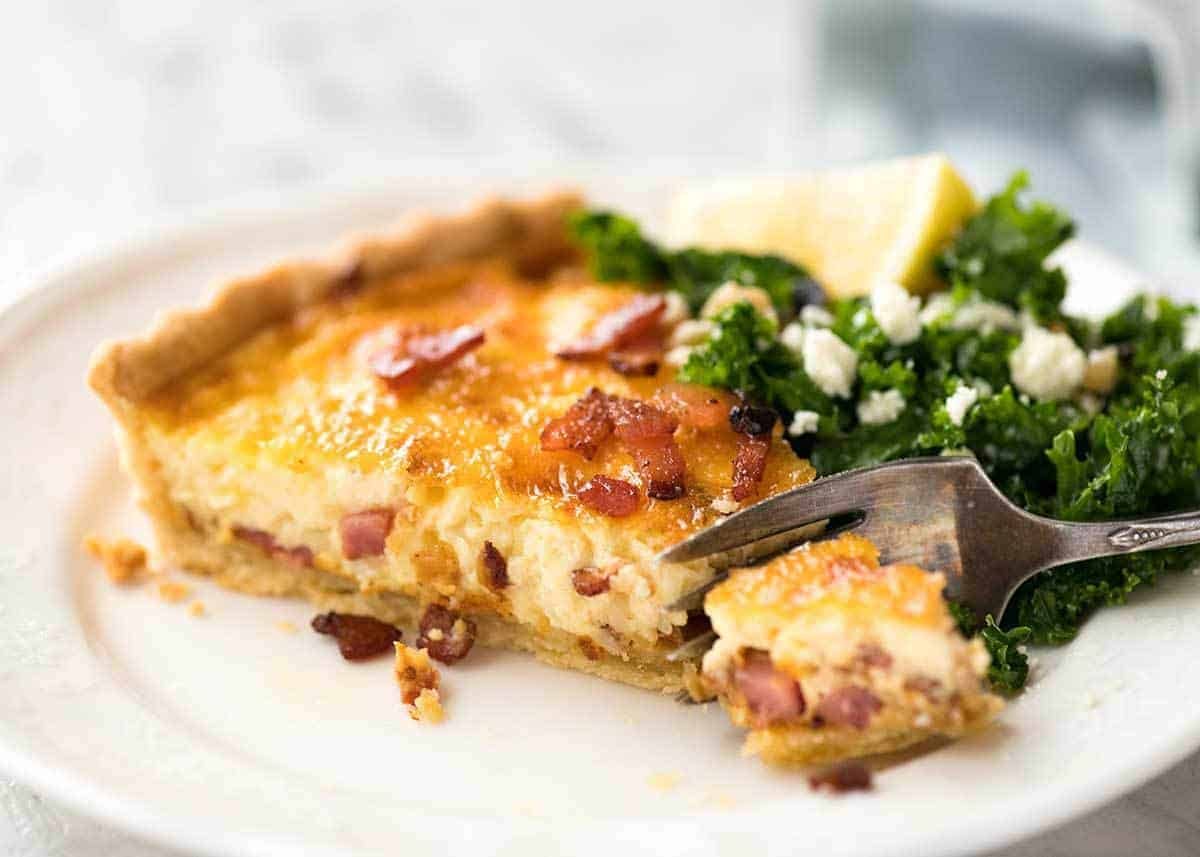 Slice of quiche lorraine on a white plate with a fork cutting into it