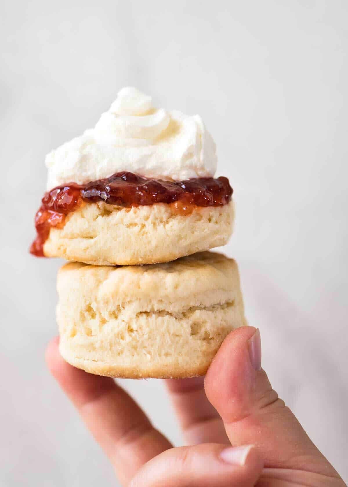 Close up of a plain scone with jam and cream being held up high, stacked on top of another scone.